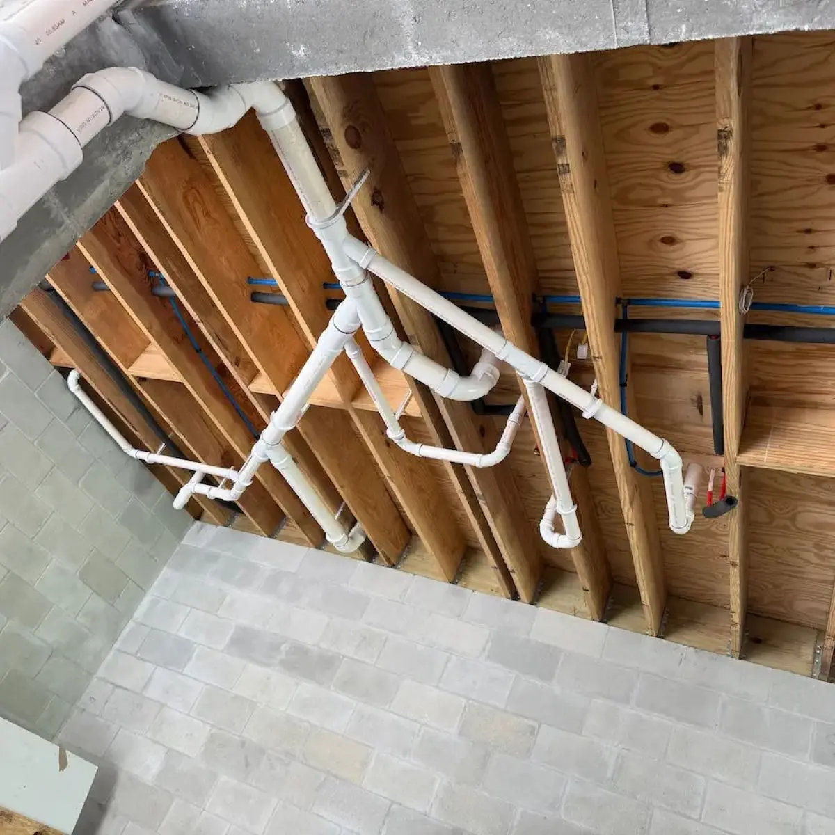 Overhead view of white PVC commercial drainage pipes secured to wood floor joists above concrete block walls.