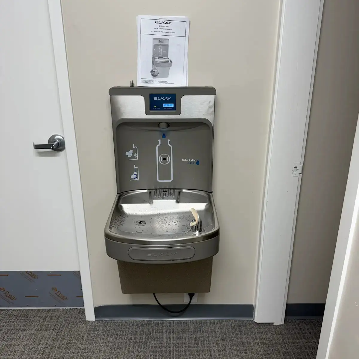 A stainless steel Elkay water bottle filling station and drinking fountain unit mounted on an office wall above gray carpeting.