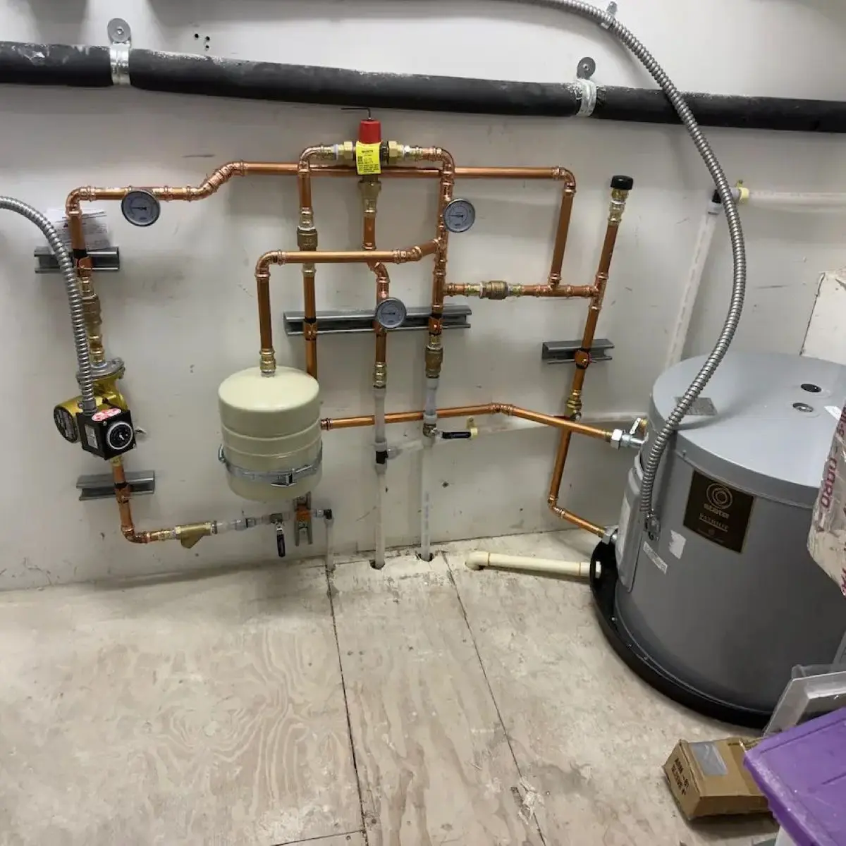 A grey State Patriot water heater tank next to a complex layout of copper pipes mounted to a wall, featuring pressure gauges and a beige thermal expansion tank.